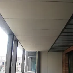 Wykończony balkon z sufitem z paneli w odcieniach beżu, widok z dołu na konstrukcję z metalowymi słupkami i widok na budynek mieszkalny w tle.