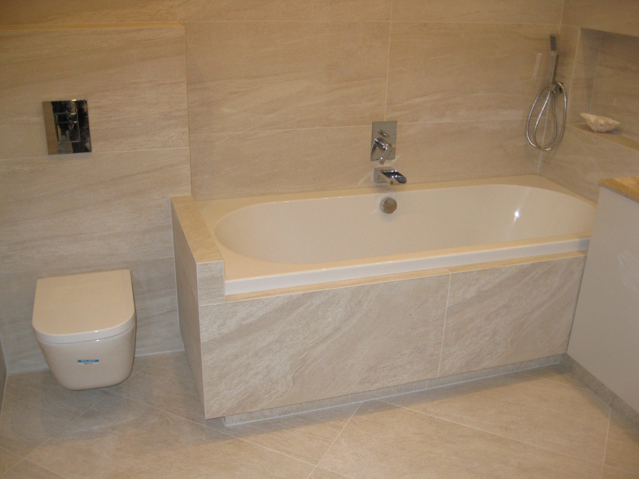 Nowoczesna łazienka z beżowymi płytkami imitującymi kamień, zabudowana wanna z chromowaną armaturą, wisząca muszla klozetowa z przyciskiem spłukującym w kolorze chromu oraz słuchawka prysznicowa.