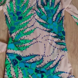 Beżowy trykot gimnastyczny z zielonymi i turkusowymi aplikacjami w kształcie liści i błyszczącymi dżetami, leżący na drewnianej podłodze.