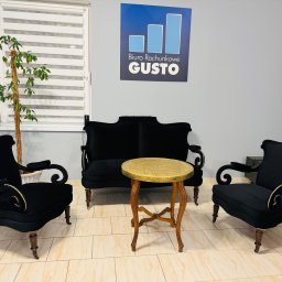 Biuro Rachunkowe Gusto Sp. z o.o. - Eleganckie wnętrze biura rachunkowego: czarne fotele i kanapa w stylu retro, złoty stolik, roślina doniczkowa i logo firmy na ścianie. Przytulna strefa dla klientów.