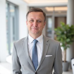 Biuro Rachunkowe Gusto Sp. z o.o. - Portret biznesowy uśmiechniętego mężczyzny w szarym garniturze i błękitnym krawacie, na jasnym tle z rośliną doniczkową i oknami.