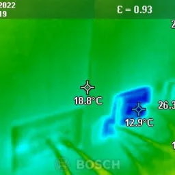 Termogram ściany z widocznymi mostkami termicznymi wokół gniazdka elektrycznego, wykonany kamerą termowizyjną Bosch, z zaznaczonymi temperaturami w różnych punktach.