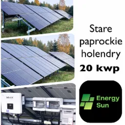 Rząd modułów fotowoltaicznych na metalowej konstrukcji, widoczny inwerter Solax Power i skrzynki zabezpieczeń, tło stanowią drzewa w jesiennej scenerii.