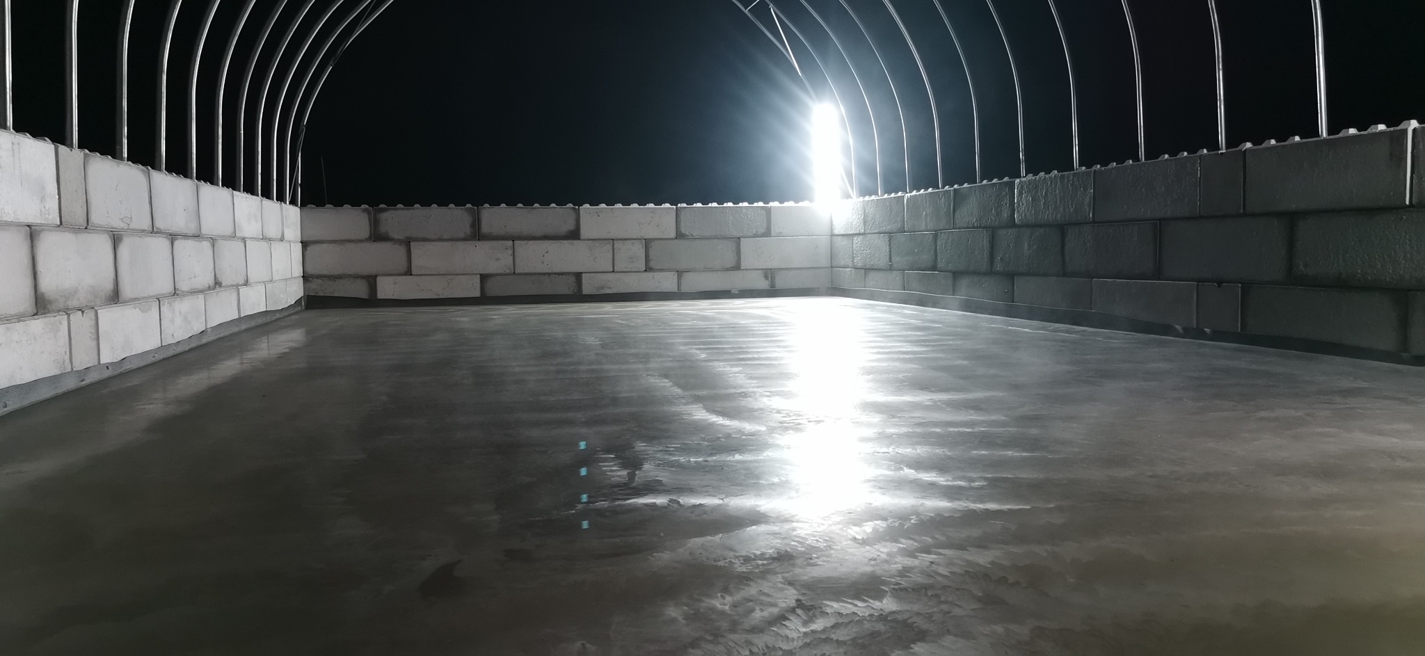 Gładka, szara posadzka w hali przemysłowej z białymi ścianami z bloczków i metalową konstrukcją dachu, oświetlona jasnym światłem.