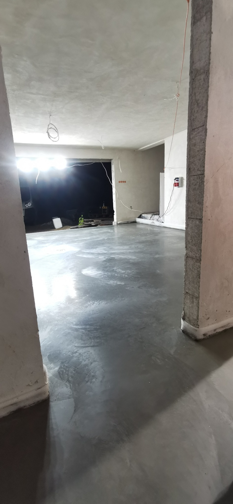 Świeżo wylana, gładka posadzka w szarym odcieniu, widok przez surową futrynę, w tle sprzęt budowlany i reflektory oświetlające wnętrze.