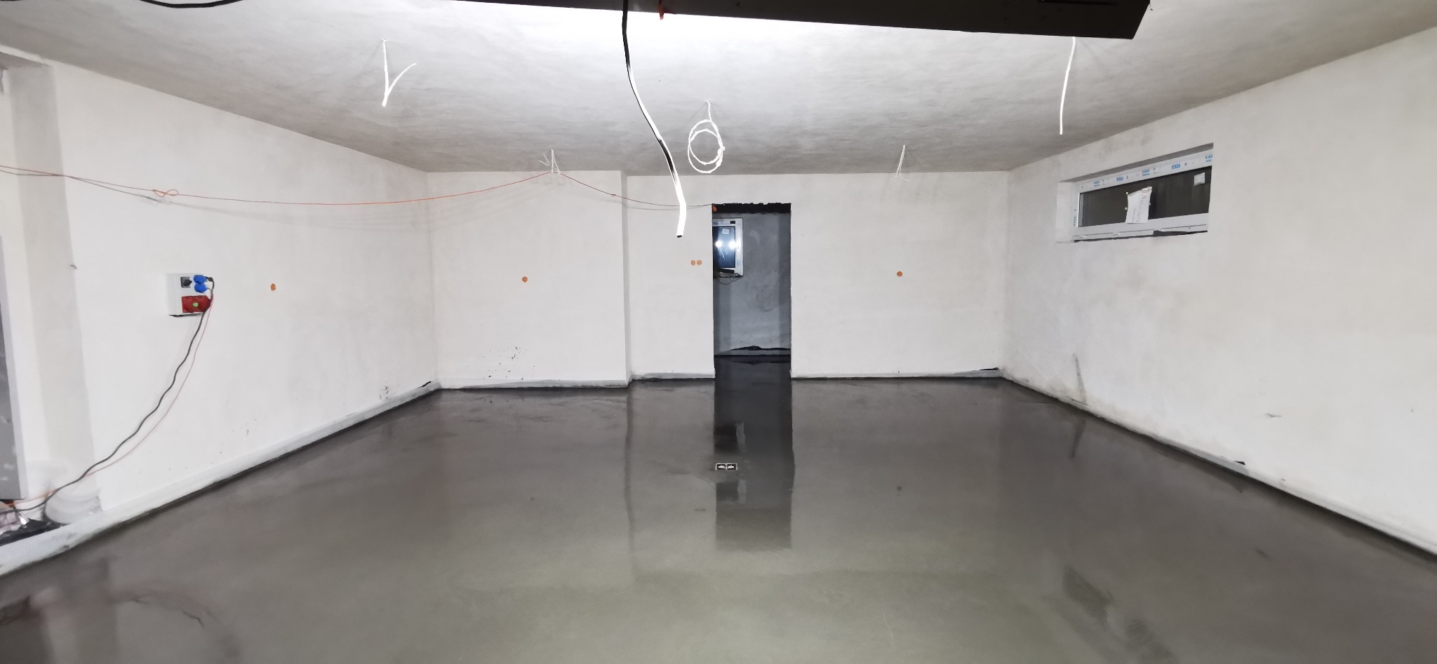 Świeżo wylana, gładka posadzka przemysłowa z widocznymi instalacjami elektrycznymi na ścianach i suficie, odblaskowa powierzchnia. Na ścianie widoczne gniazdka elektryczne.