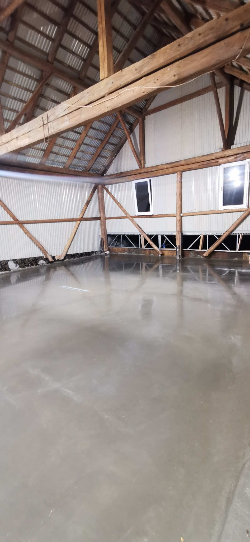 Świeżo wylana, gładka posadzka w hali z drewnianą konstrukcją dachu i ścianami z blachy falistej.
