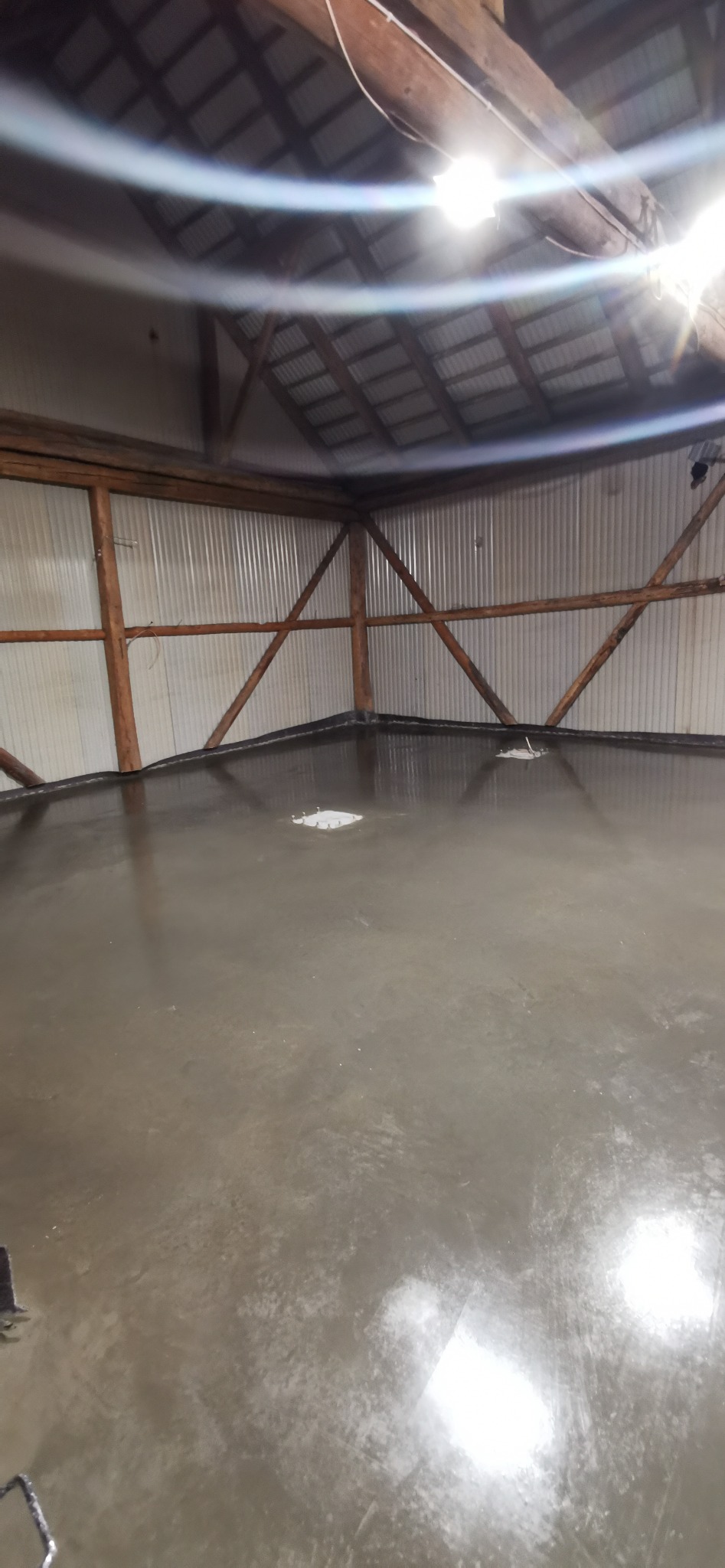 Świeżo wylana, gładka posadzka w hali przemysłowej z drewnianą konstrukcją dachu i ścianami z blachy falistej, widoczne punkty świetlne od lamp.