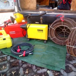 Sprzęt hydrauliczny REMS i RIDGID do czyszczenia rur, w tym spirale, urządzenia elektryczne i skrzynka narzędziowa, rozłożony na zielonej plandece przed otwartym bagażnikiem samochodu dostawczego.