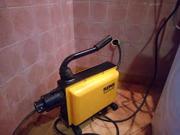 Żółta, elektryczna przepychaczka do rur REMS w rogu łazienki z różowymi płytkami, kabel zasilający zwinięty obok urządzenia.