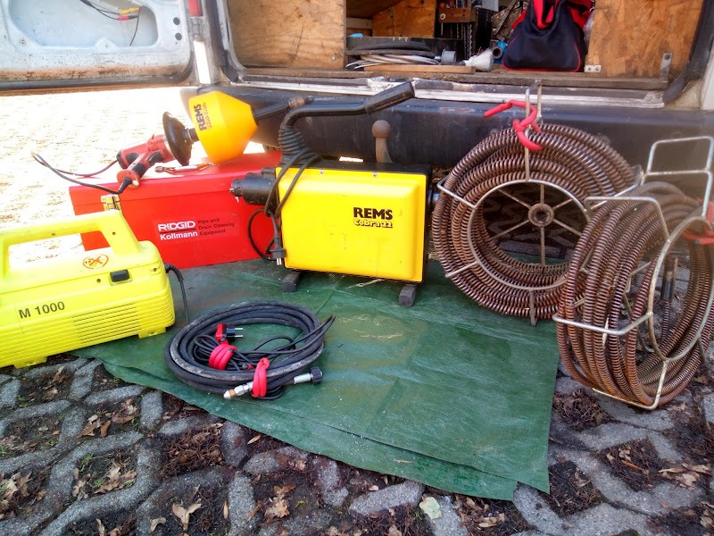 Sprzęt hydrauliczny REMS i RIDGID do czyszczenia rur, w tym spirale, urządzenia elektryczne i skrzynka narzędziowa, rozłożony na zielonej plandece przed otwartym bagażnikiem samochodu dostawczego.