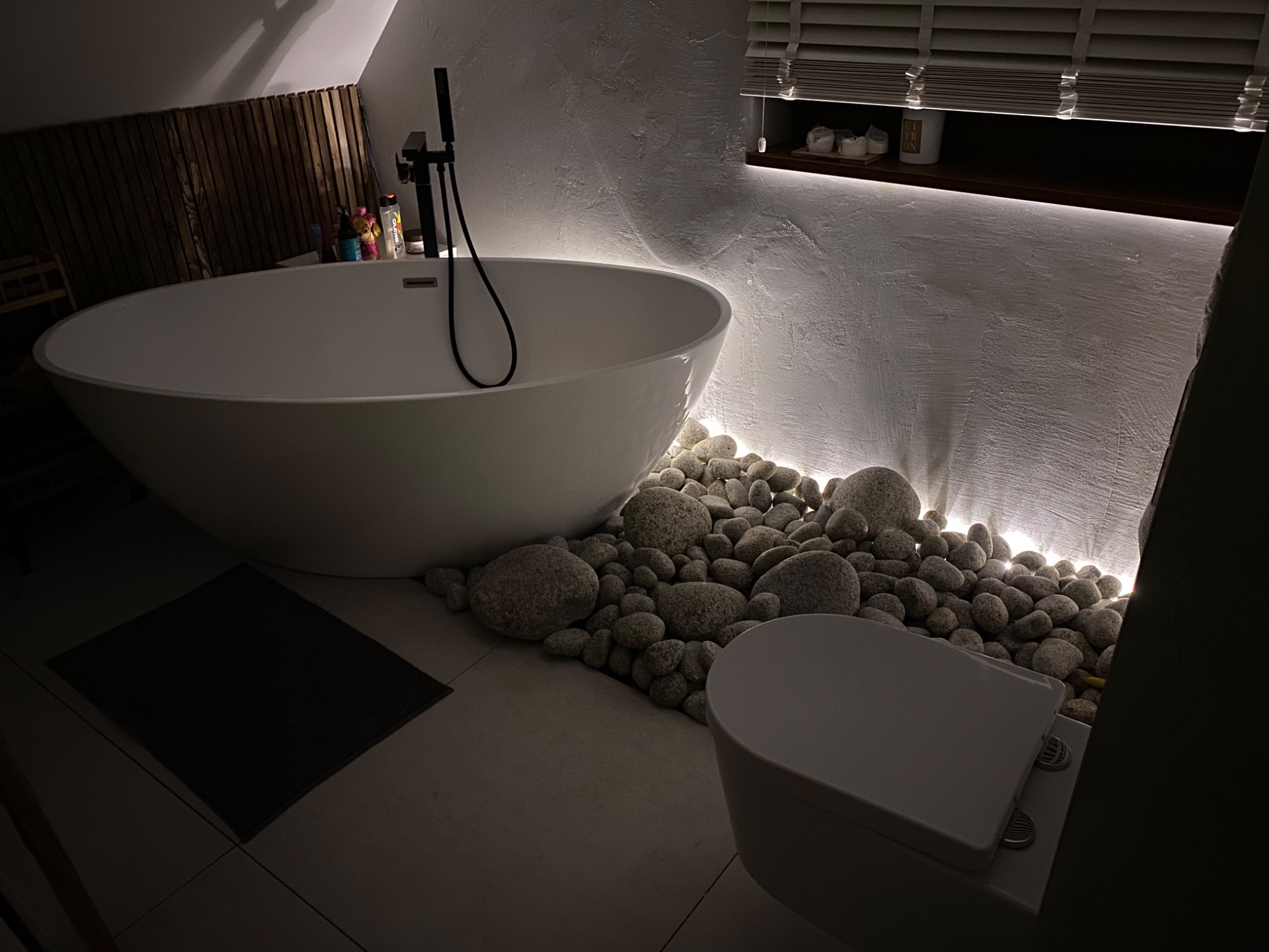 Nowoczesna łazienka w stylu minimalistycznym z wolnostojącą białą wanną, czarną armaturą, dekoracyjnymi kamieniami z podświetleniem LED, płytkami podłogowymi i drewnianymi żaluzjami w oknie.