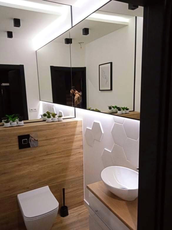 Nowoczesna łazienka z drewnianymi panelami, heksagonalnymi płytkami 3D i podświetlanym lustrem, widok na toaletę i umywalkę.