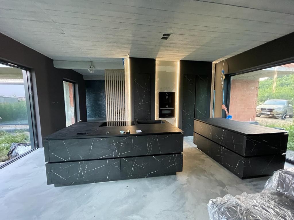 Nowoczesna kuchnia w trakcie wykańczania, z czarnymi szafkami z marmurkowym wzorem, surowym betonowym sufitem i dużymi oknami z widokiem na zewnątrz, widoczne elementy wykończeniowe i folie ochronne.