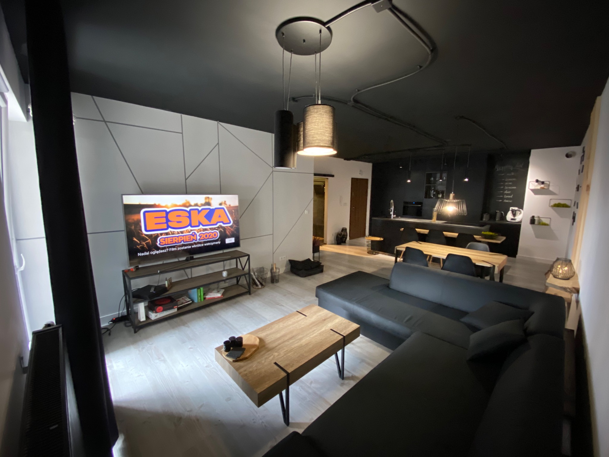 Nowoczesny salon z czarnym sufitem, geometryczną ścianą, drewnianym stolikiem kawowym i narożną sofą, widok na otwartą kuchnię z drewnianym blatem i czarnymi szafkami.