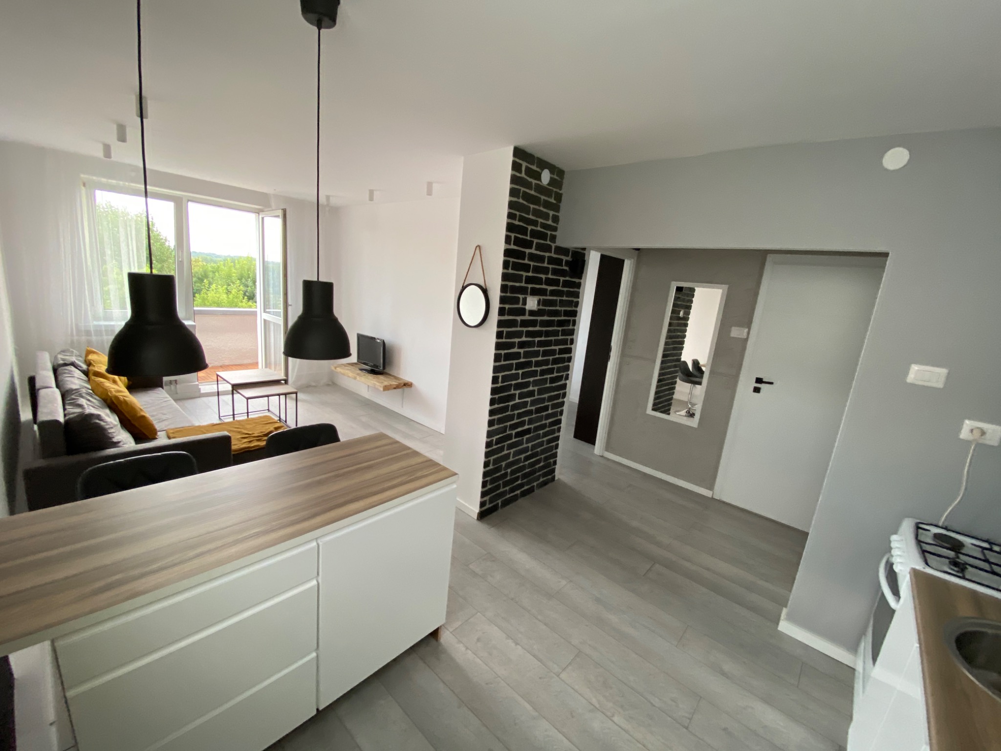 Nowoczesne wnętrze mieszkania z szarą podłogą, białymi ścianami, fragmentem ściany z czarnej cegły, dwoma czarnymi lampami wiszącymi, szarą sofą z żółtymi poduszkami i widokiem na zieleń za oknem.