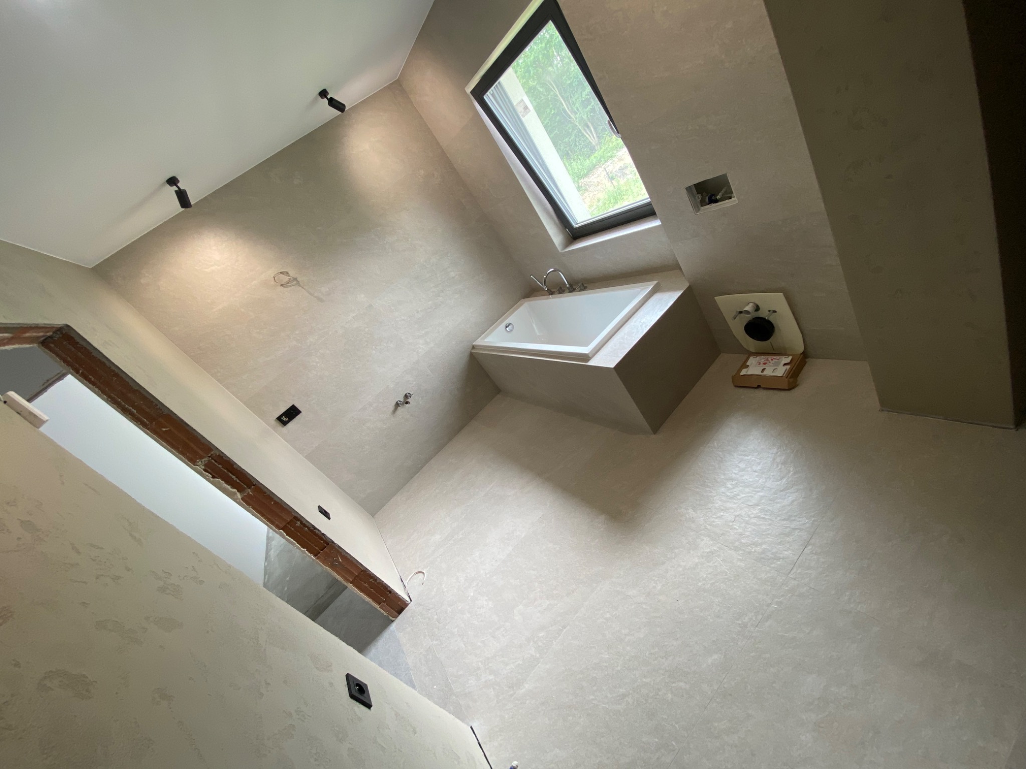 Nowoczesna łazienka w trakcie wykończenia z surowymi ścianami w odcieniach szarości, biała wanna pod oknem dachowym i widoczne punkty oświetleniowe na suficie.