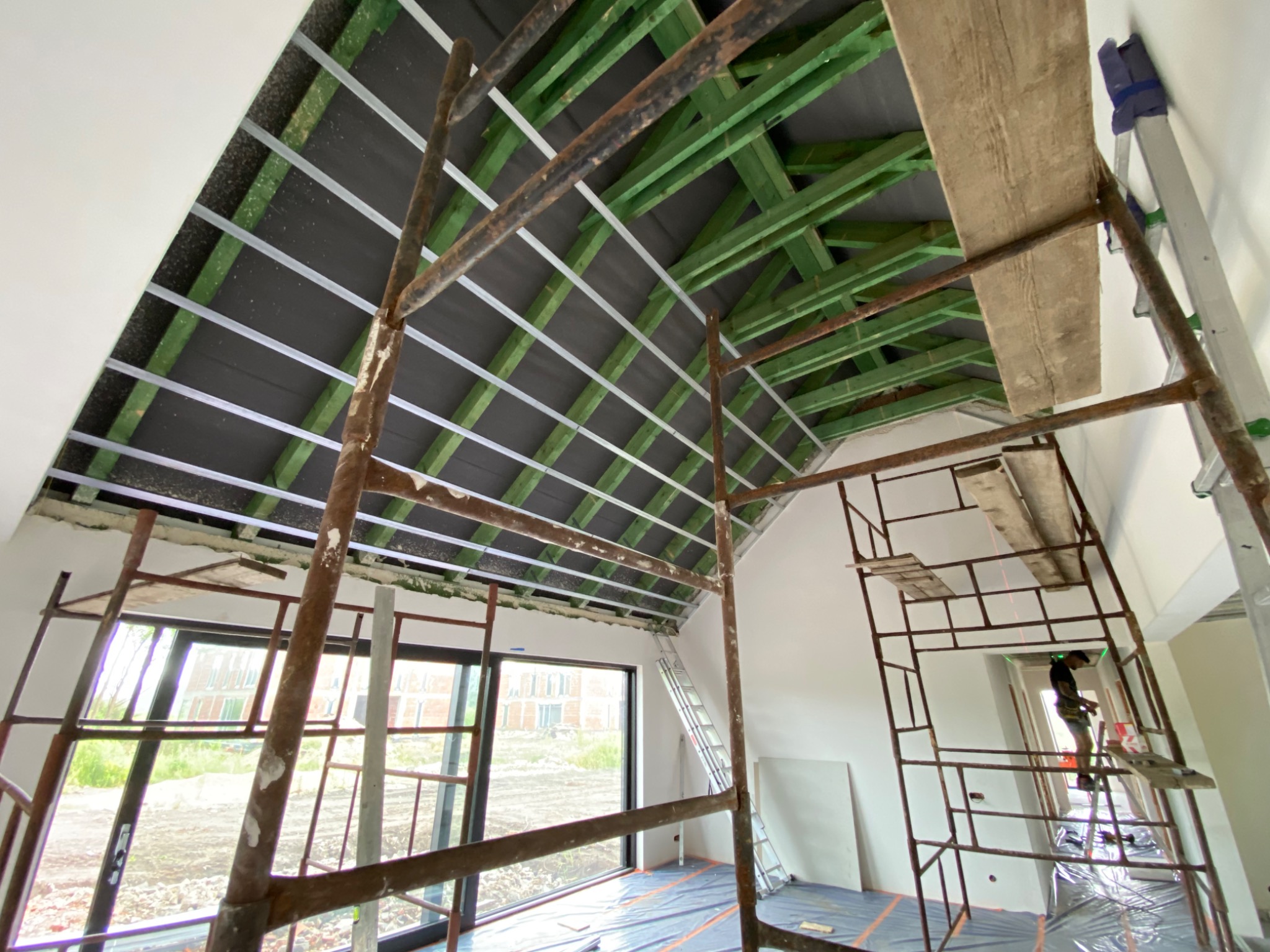 Ocieplanie poddasza w trakcie realizacji: widoczne rusztowania, zielone belki konstrukcyjne, czarna warstwa izolacyjna oraz metalowe profile mocujące.