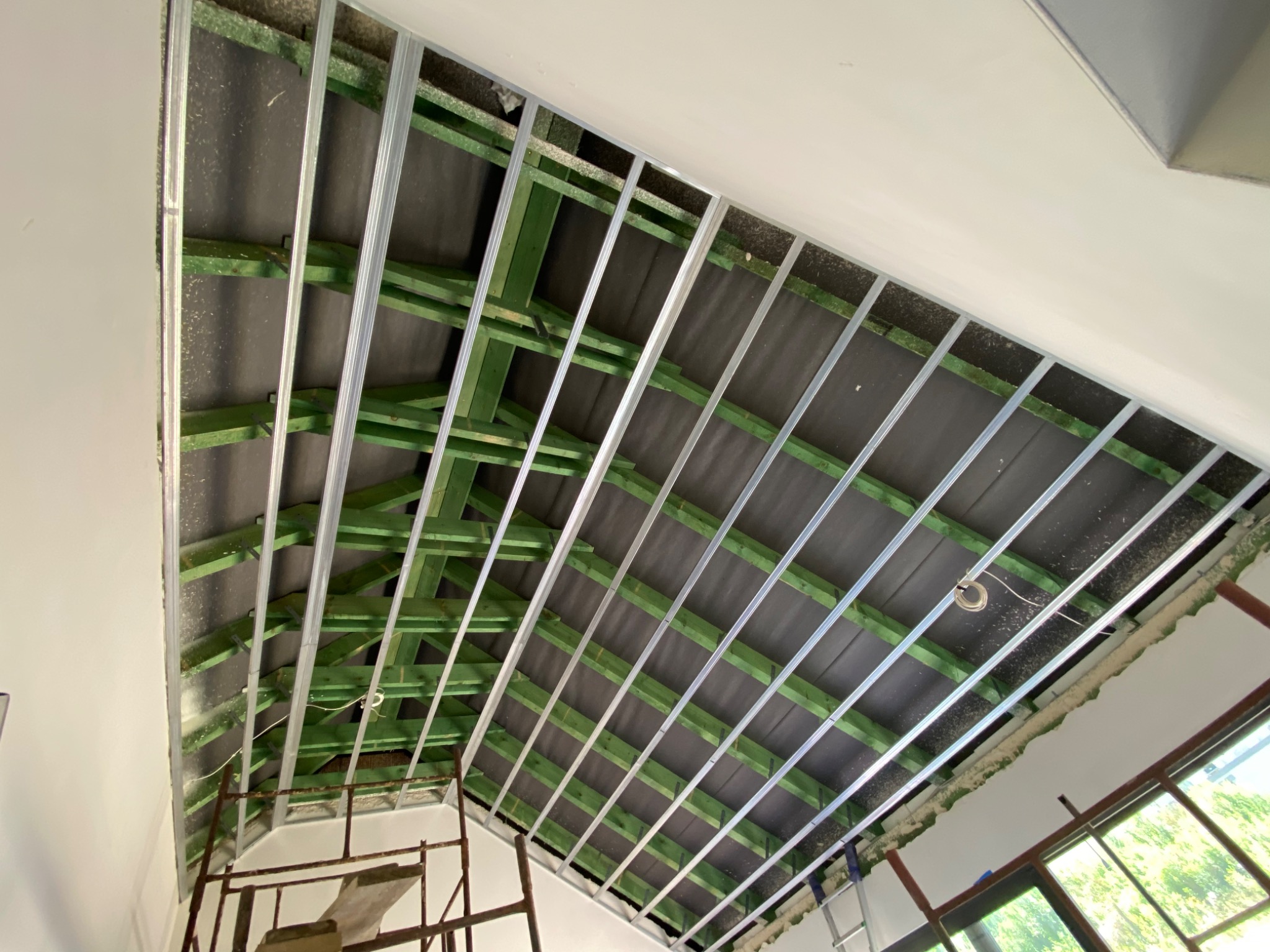 Konstrukcja podwieszanego sufitu z zielonych belek drewnianych i metalowych profili, widok od dołu, czarna mata izolacyjna widoczna między elementami konstrukcji, fragment rusztowania w lewym...