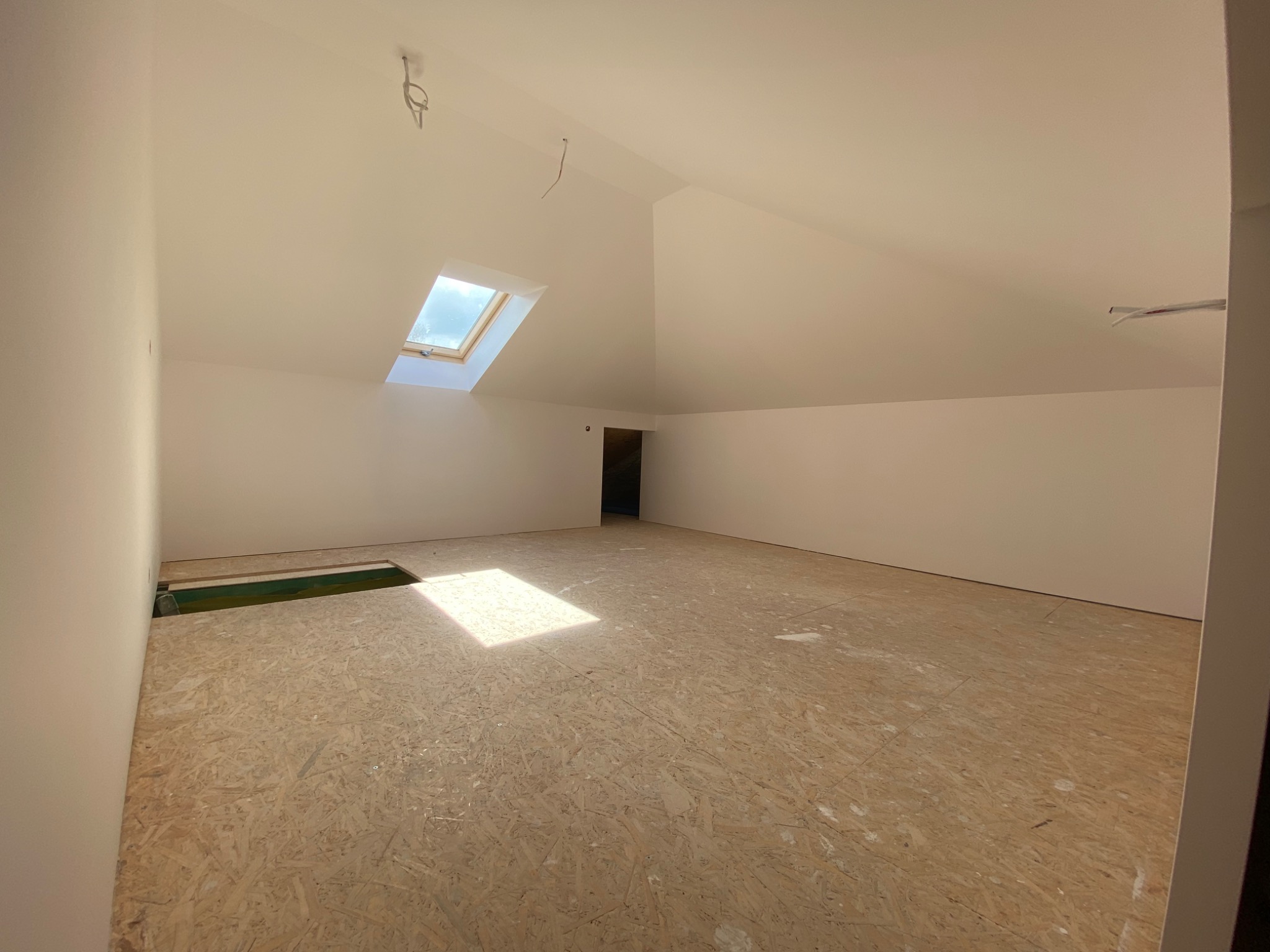 Poddasze z oknem dachowym, świeżo pomalowane ściany na biało, podłoga wyłożona płytami OSB, widoczne kable wystające z sufitu i otwór w podłodze z zielonymi elementami.