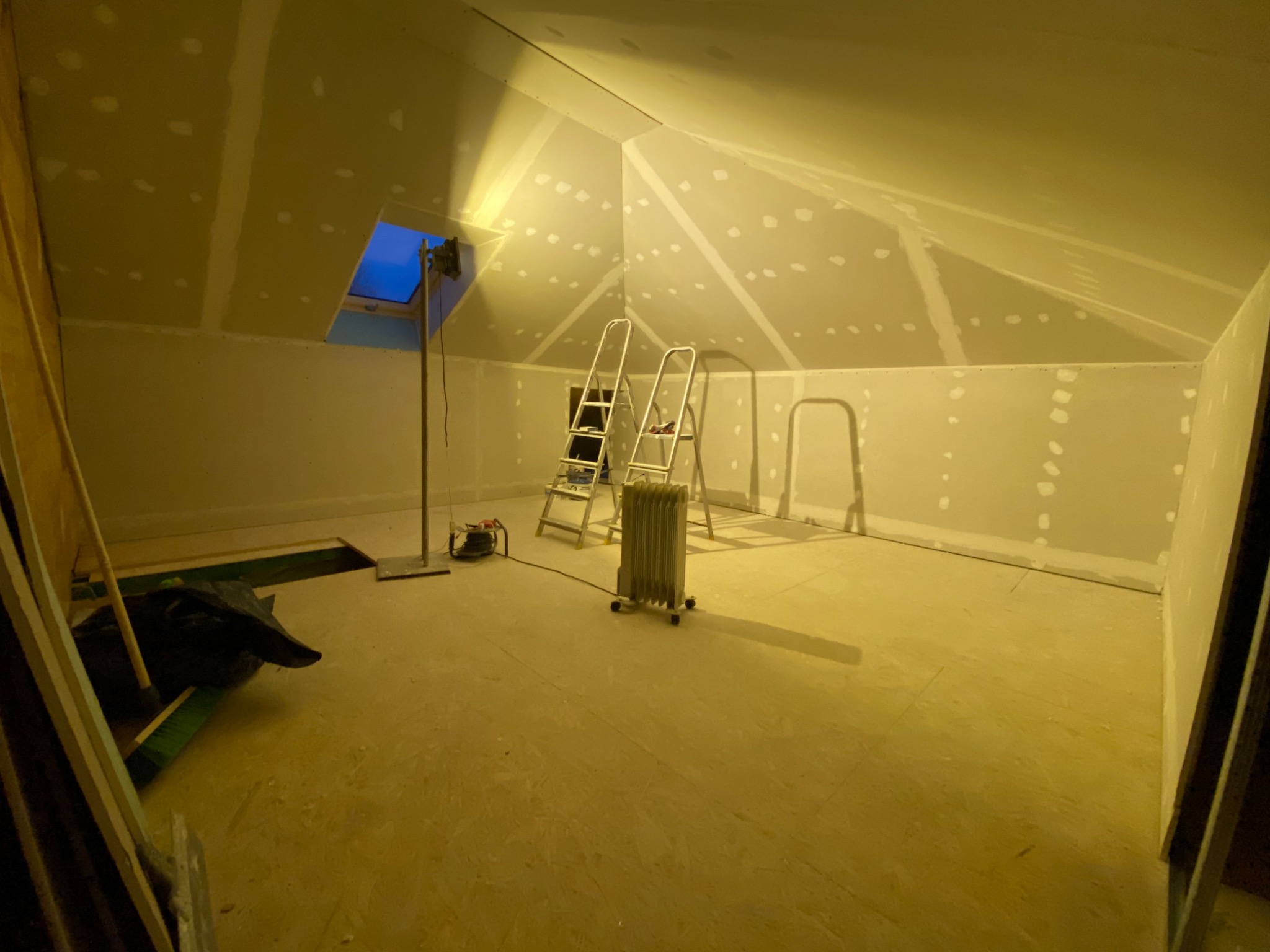 Poddasze w trakcie ocieplania, widoczne płyty kartonowo-gipsowe z widocznymi łączeniami, drabina, grzejnik elektryczny i lampa doświetlająca