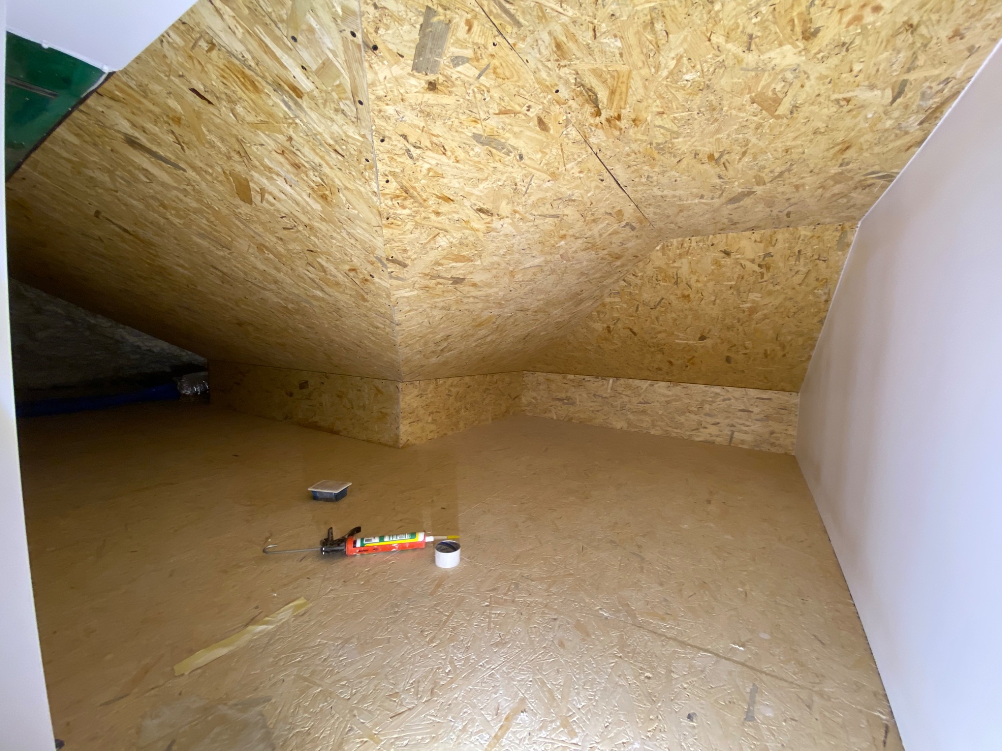 Poddasze wyłożone płytami OSB, widoczny pistolet do silikonu, taśma malarska i pojemnik na podłodze, ściany pomalowane na biało.