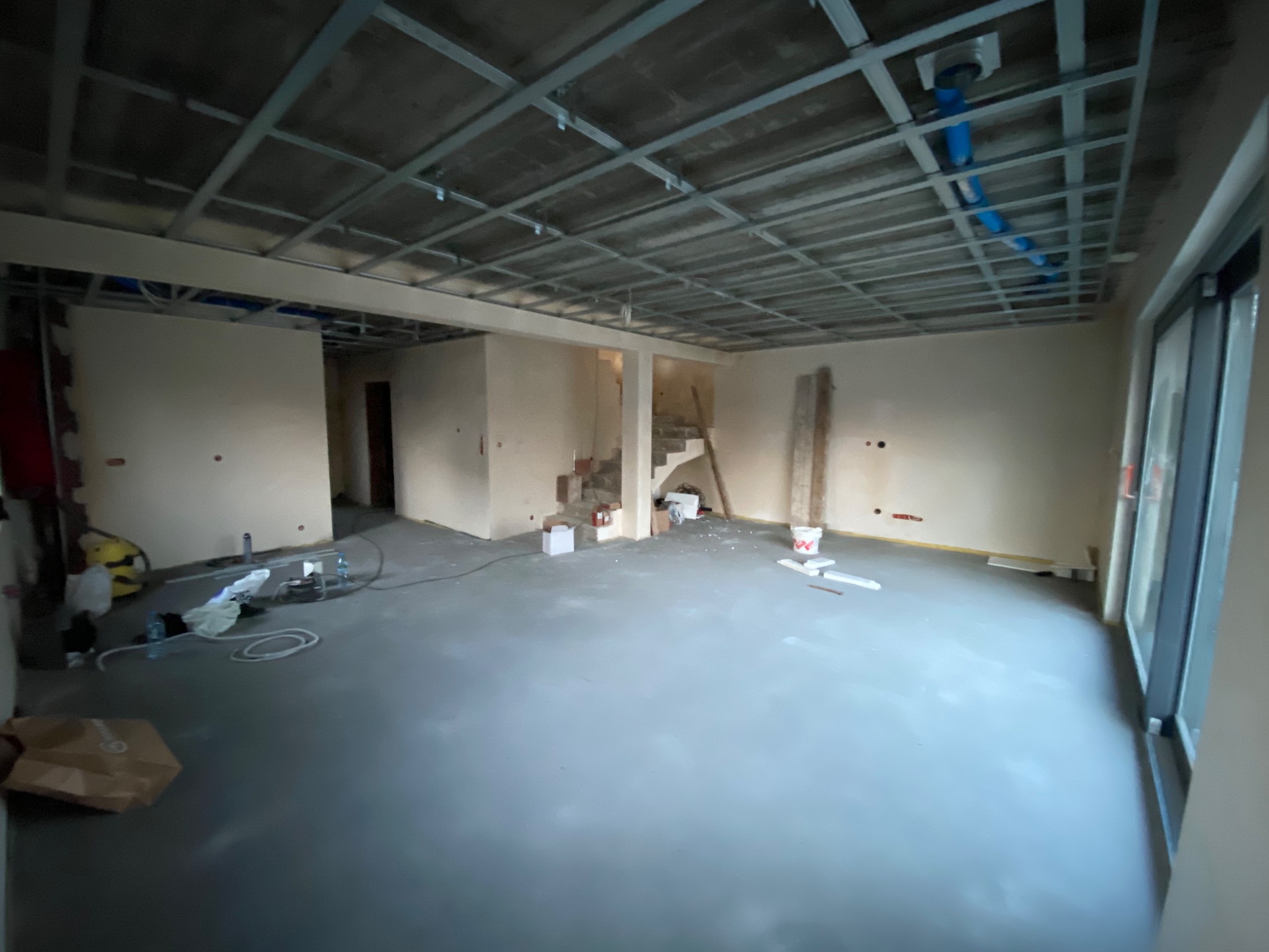 Wnętrze w trakcie prac wykończeniowych: surowy strop podwieszany z widocznymi instalacjami, wylewka podłogowa, ściany przygotowane do malowania, widoczne schody w głębi pomieszczenia.
