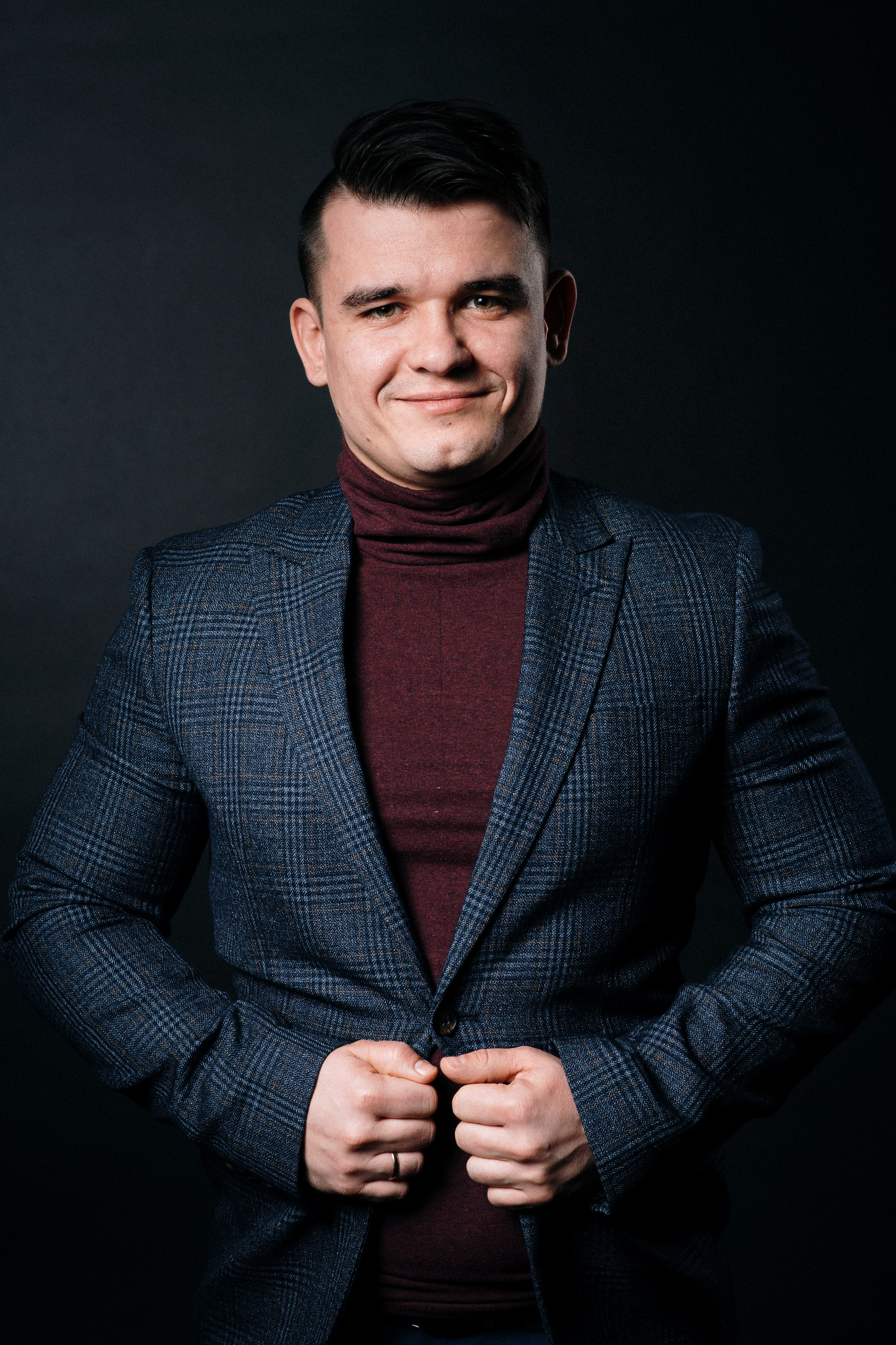 Uśmiechnięty mężczyzna w bordowym golfie i granatowej marynarce w kratę, zapinający guzik marynarki, na ciemnym tle.