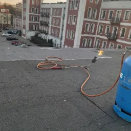 Płomień z palnika dekarskiego na płaskim, bitumicznym dachu, w tle budynek mieszkalny z czerwonej cegły.