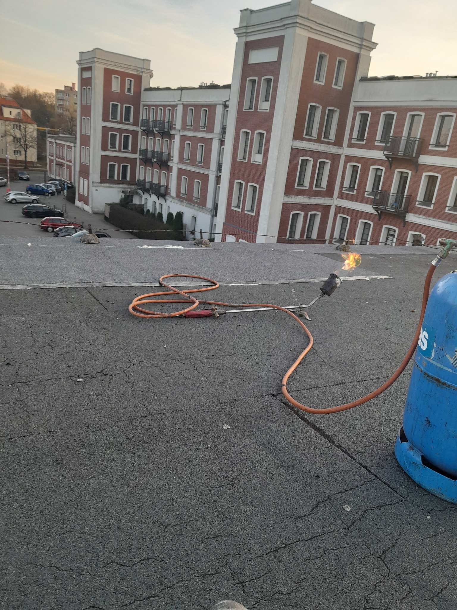 Płomień z palnika dekarskiego na płaskim, bitumicznym dachu, w tle budynek mieszkalny z czerwonej cegły.