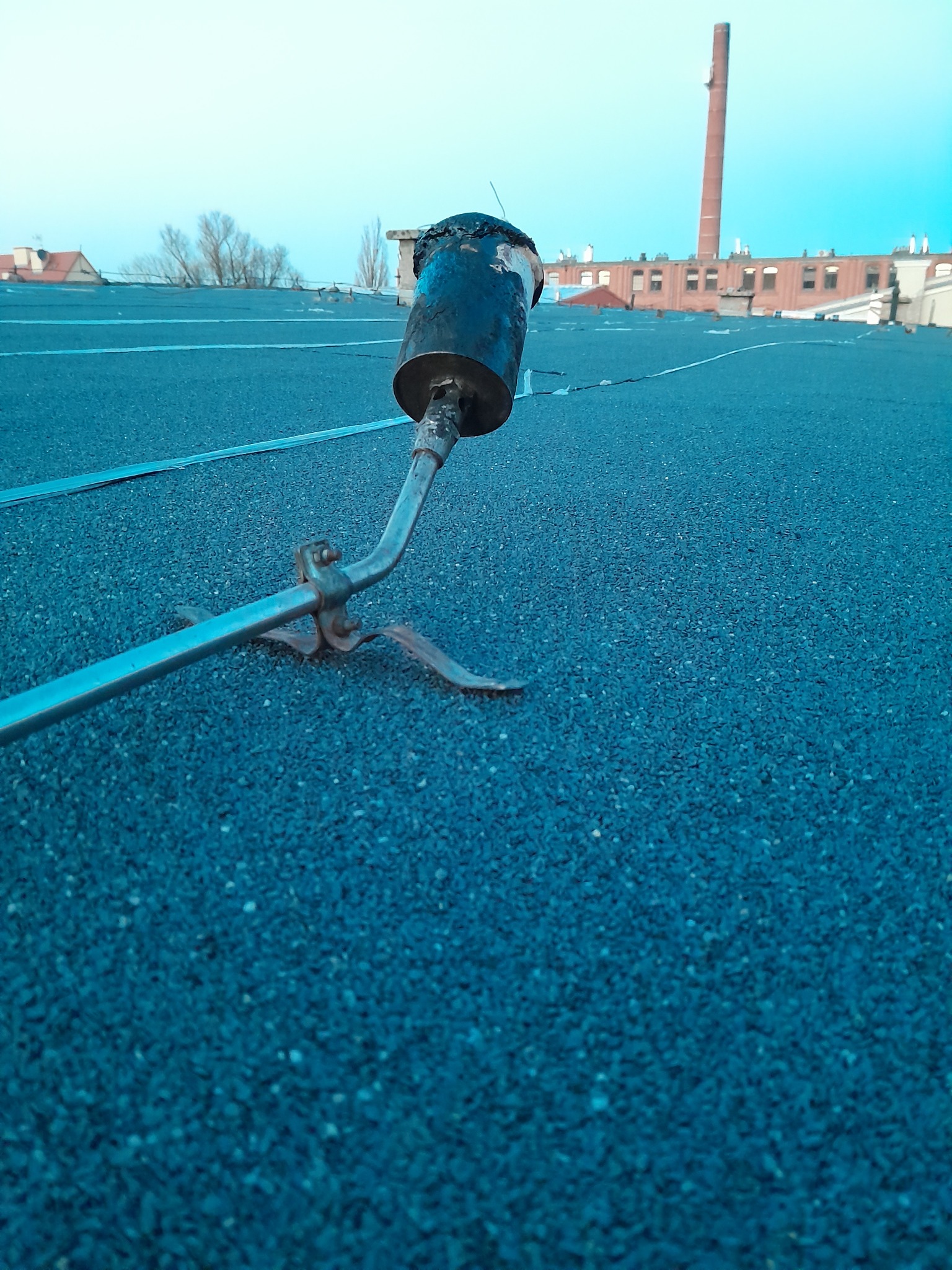 Palnik dekarski na pokrytym papą dachu w trakcie pracy, w tle ceglany budynek z kominem.