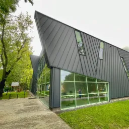 Nowoczesny budynek z ciemną elewacją i dużymi szklanymi oknami, otoczony zielenią, widok z zewnątrz na chodnik z kostki.