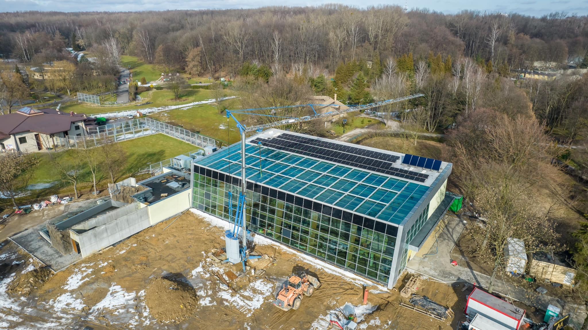 Budynek z przeszkloną fasadą i panelami słonecznymi na dachu, w trakcie budowy, widok z góry, z dźwigiem i sprzętem budowlanym na placu budowy.