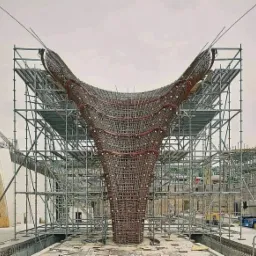 Skomplikowana konstrukcja z prętów zbrojeniowych w kształcie kielicha, otoczona rusztowaniami na placu budowy. Widoczne liczne poziome i pionowe pręty, tworzące gęstą siatkę. W tle budynek...