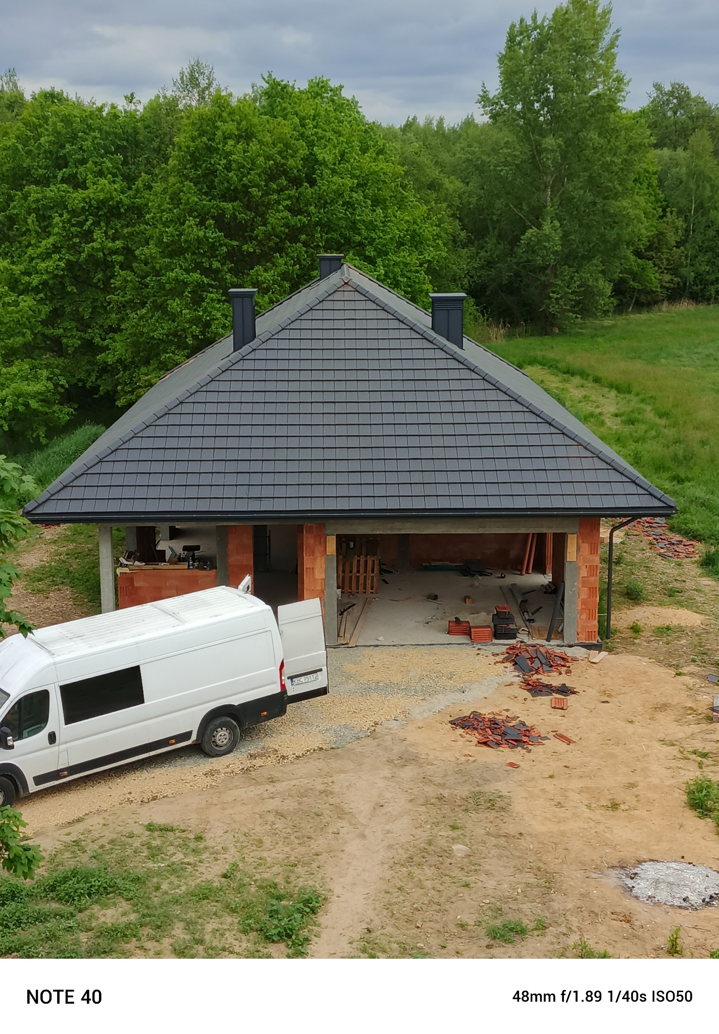 Wysoki kąt: budowa domu z ciemnym dachem z dachówki, częściowo otwartą bramą garażową i białym vanem dostawczym na żwirowym podłożu. W tle zielone drzewa.