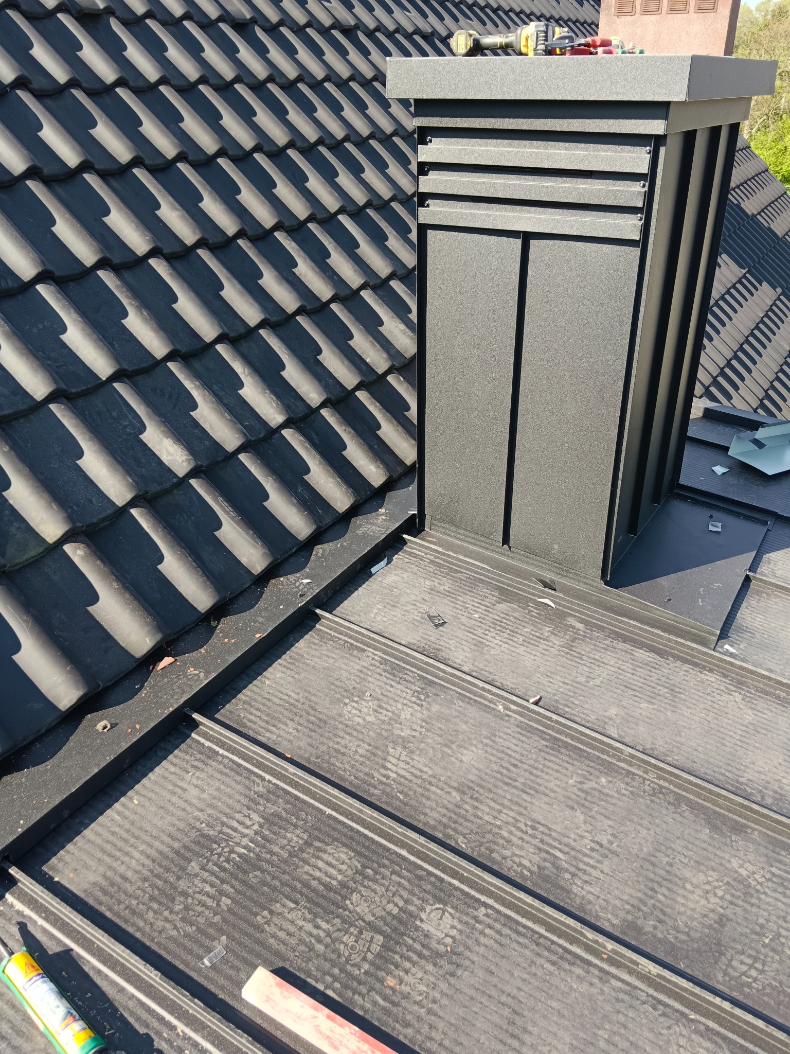 Szczegółowe ujęcie dachu z czarną dachówką i kominem w kolorze grafitowym. Widoczne narzędzia i materiały dekarskie. Ujęcie z góry, w słoneczny dzień.