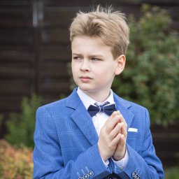 Michał Ciepielowski Dziki kadr - Portret eleganckiego chłopca w niebieskim garniturze w kratę i granatowej muszce, z rękami złożonymi w modlitewnym geście, na tle zieleni i brązowego płotu.