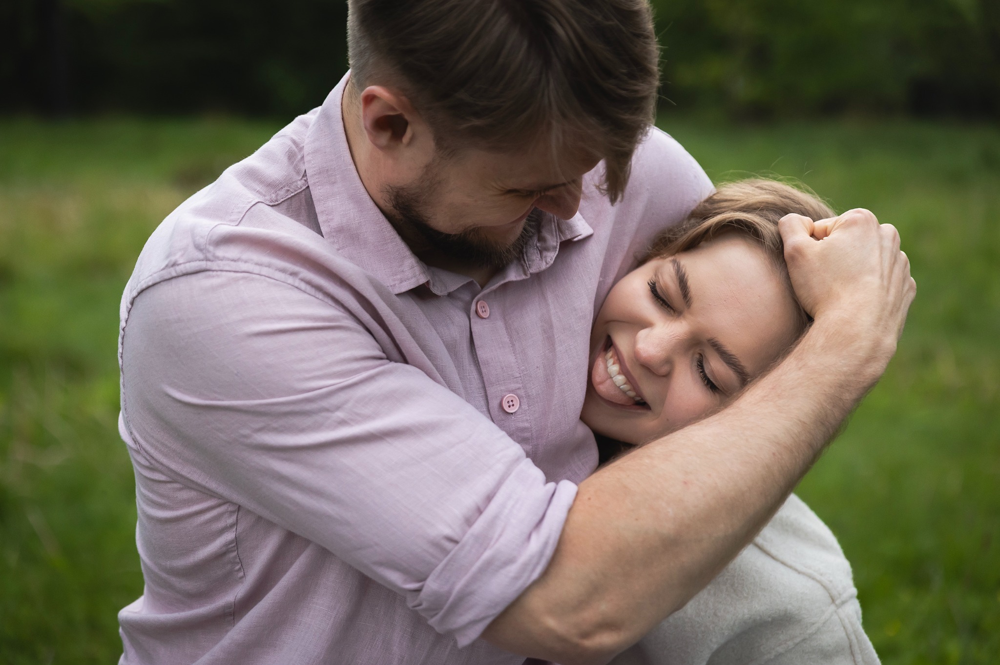 Uśmiechnięta para w objęciach na tle zieleni. Mężczyzna w różowej koszuli obejmuje kobietę, która z zamkniętymi oczami i uśmiechem opiera się o niego.