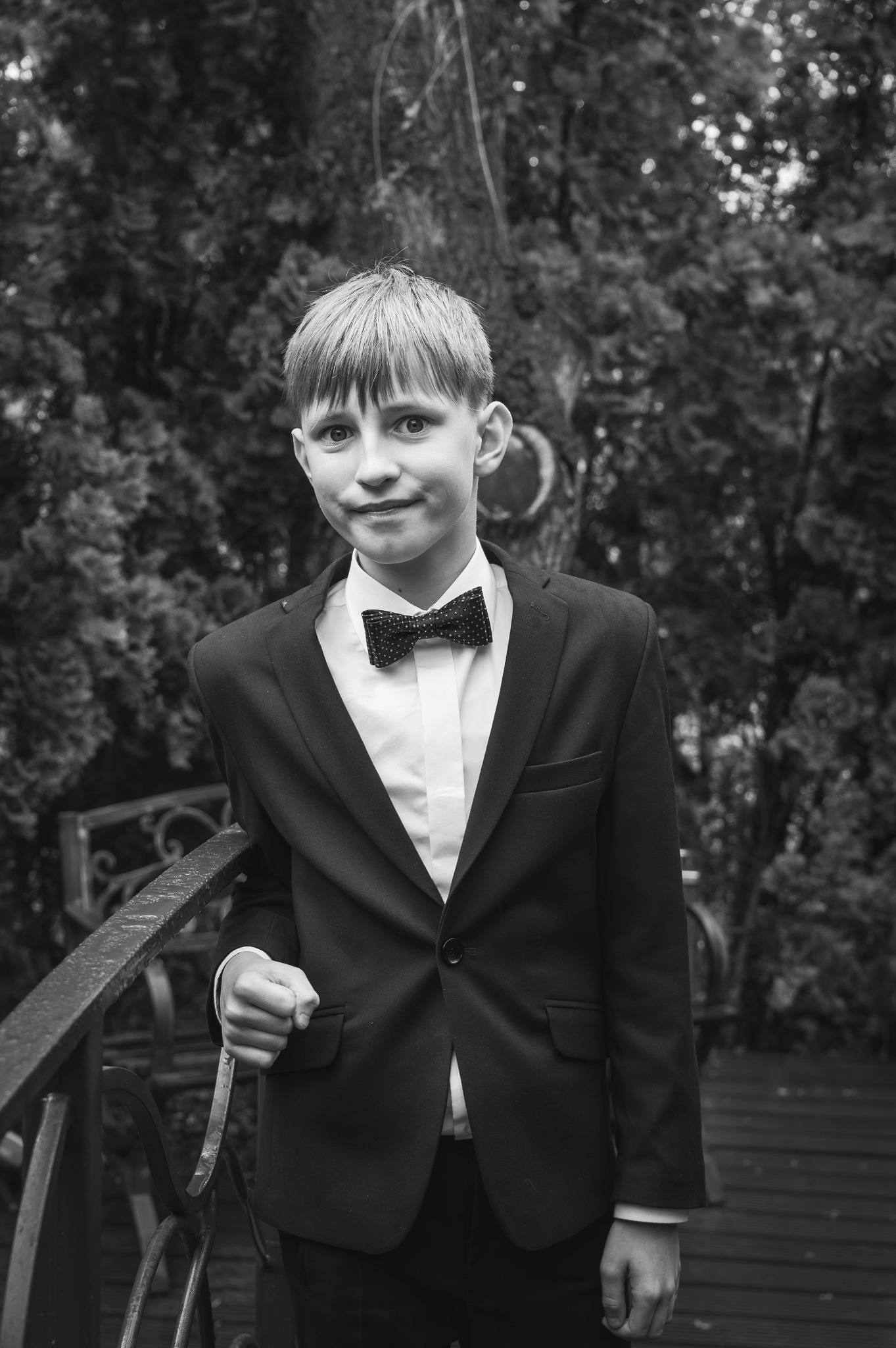 Czarno-białe zdjęcie elegancko ubranego chłopca w Łodzi, w garniturze i muszce, opierającego się o metalową barierkę na tle zieleni. Ujęcie portretowe.