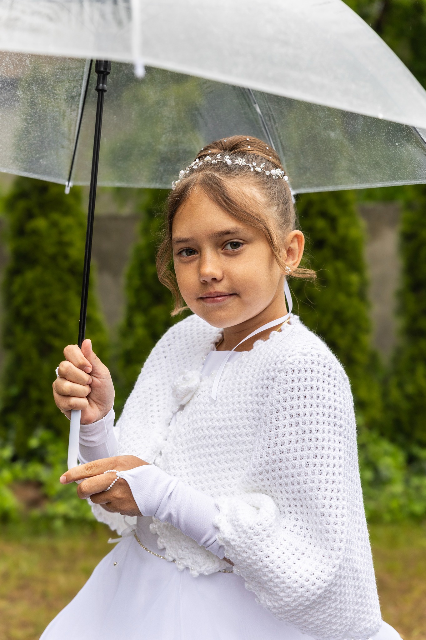 Dziewczynka w białej sukience i z przezroczystym parasolem na tle zieleni, pozująca do zdjęcia komunijnego w plenerze.
