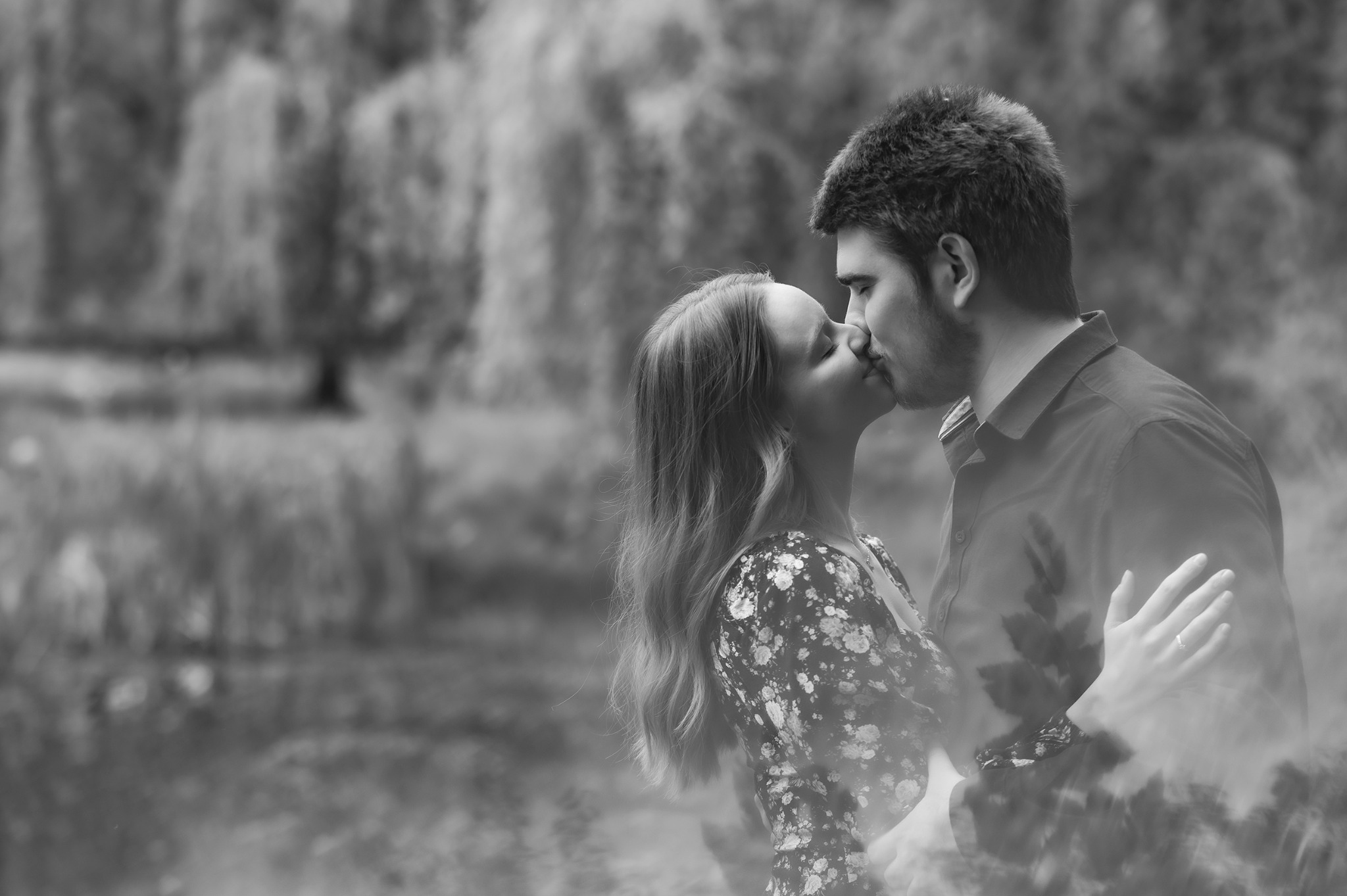Czarno-białe zdjęcie całującej się pary na tle drzew i wody. Kobieta w kwiecistej sukience, mężczyzna w koszuli. Ujęcie z profilu, delikatne światło, romantyczna atmosfera.