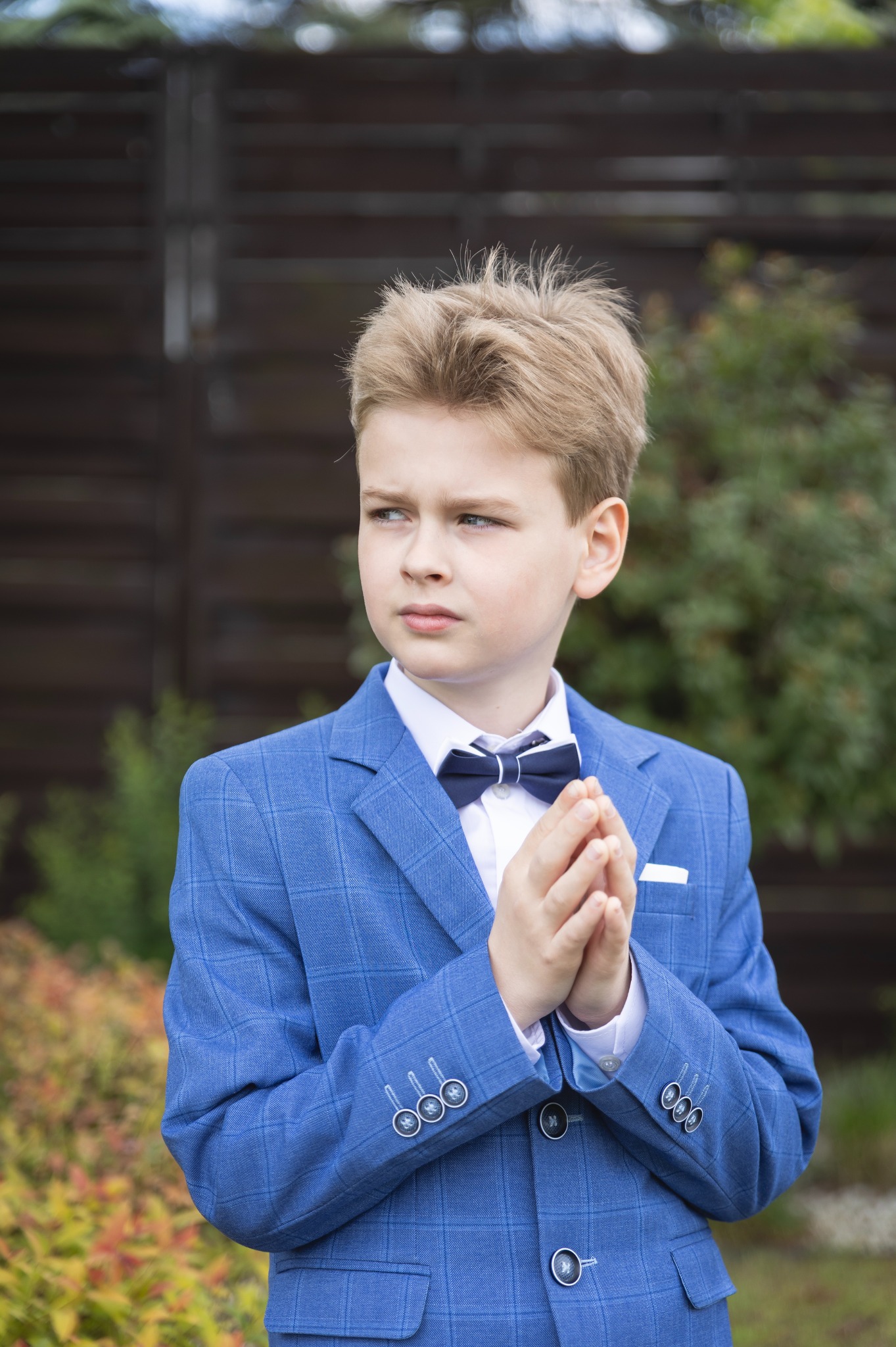 Portret eleganckiego chłopca w niebieskim garniturze w kratę i granatowej muszce, z rękami złożonymi w modlitewnym geście, na tle zieleni i brązowego płotu.