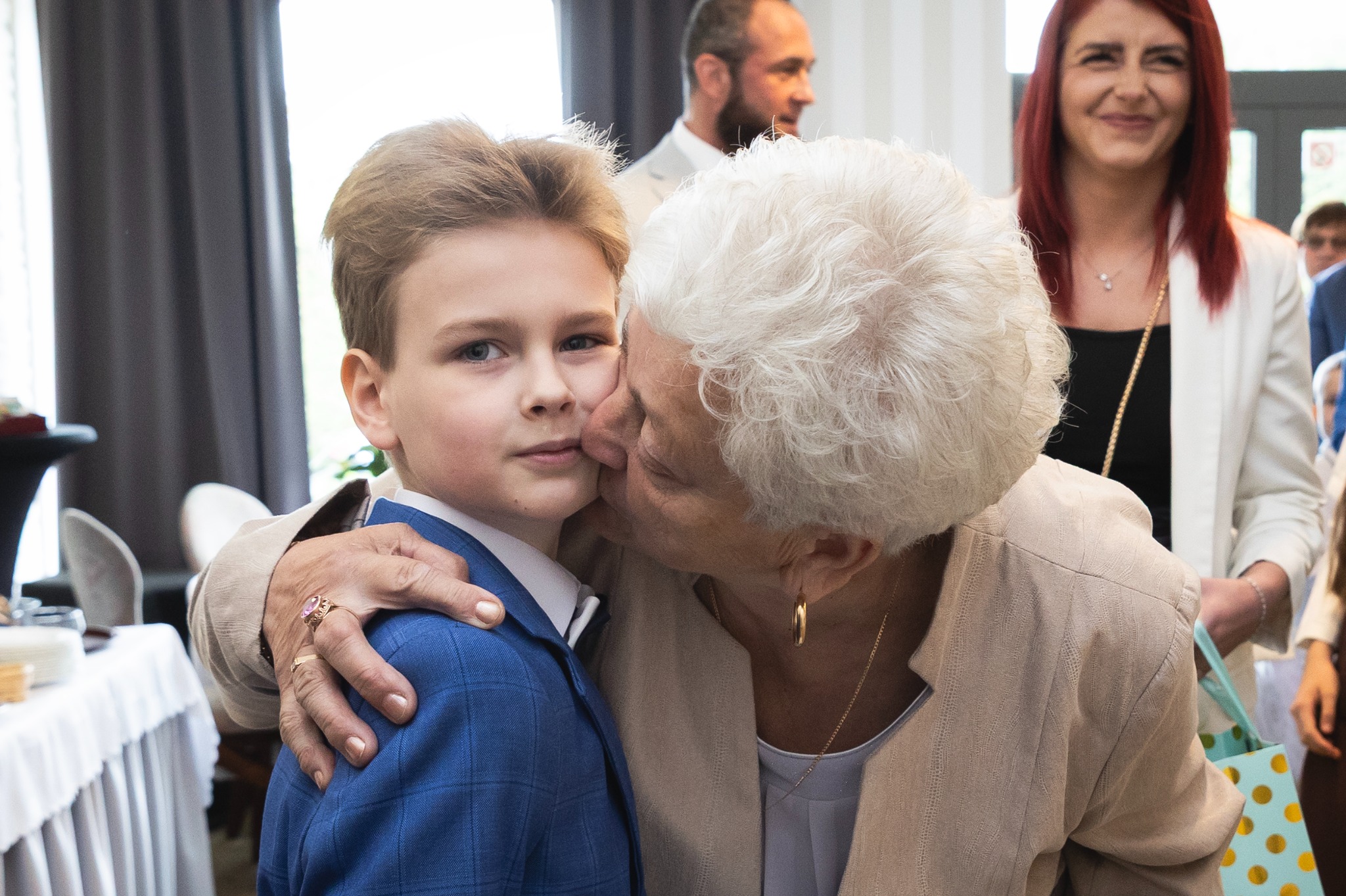 Babcia całuje wnuka w policzek podczas uroczystości rodzinnej. Chłopiec w eleganckim, niebieskim garniturze, w tle uśmiechnięte osoby. Ciepłe, naturalne światło.