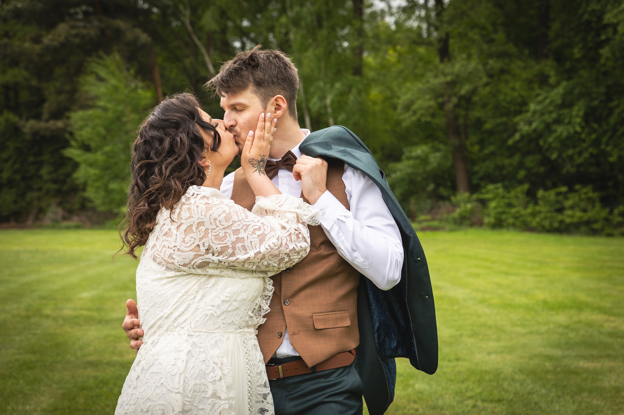 Para młoda: panna młoda w koronkowej sukni i pan młody w brązowej kamizelce, całują się na tle zielonego trawnika i drzew. Panna młoda ma tatuaż na dłoni.