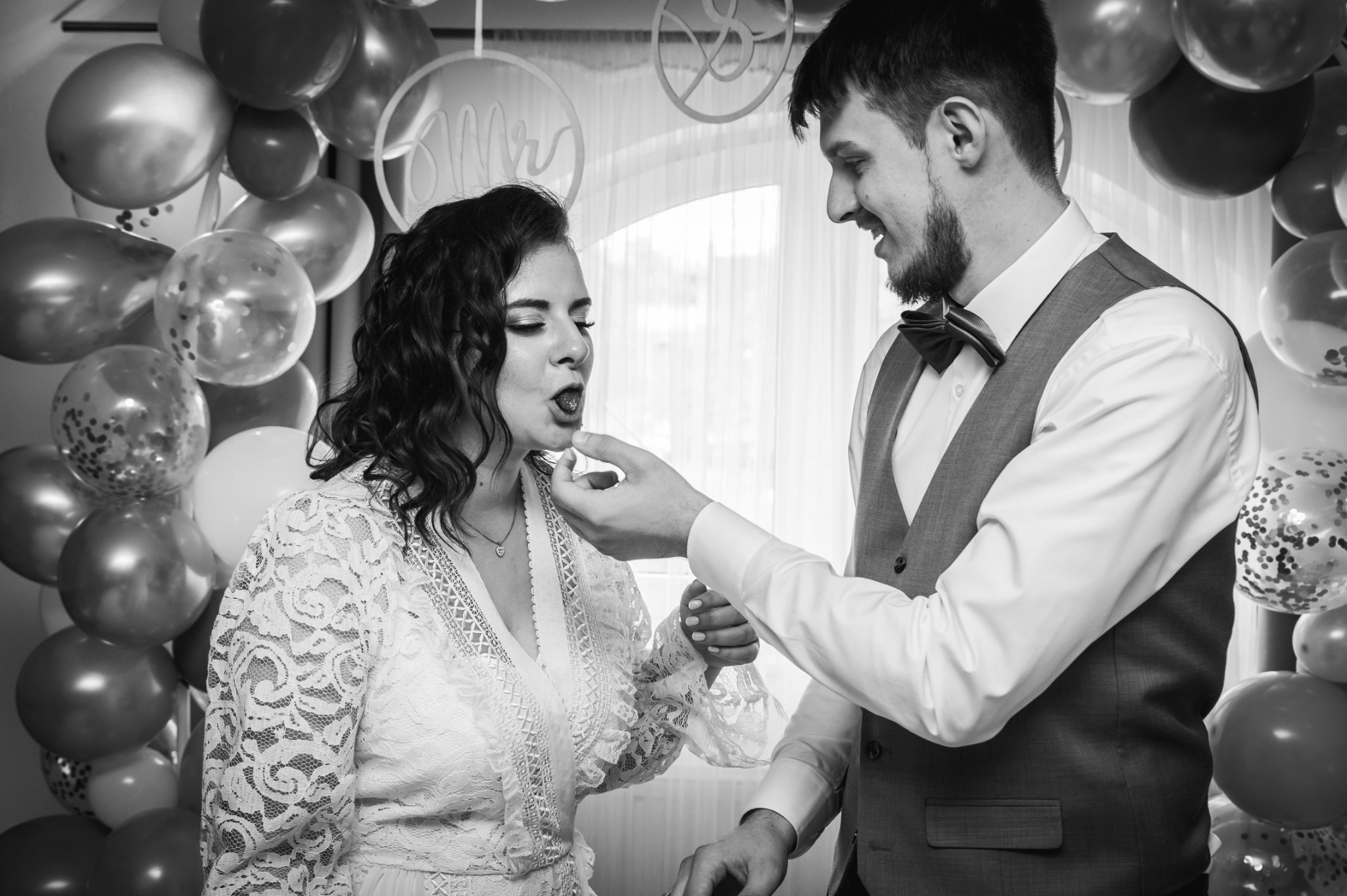 Czarno-białe zdjęcie pary młodej podczas wesela: pan młody karmi pannę młodą słodkością na tle balonów i dekoracji 'Mr & Mrs'.
