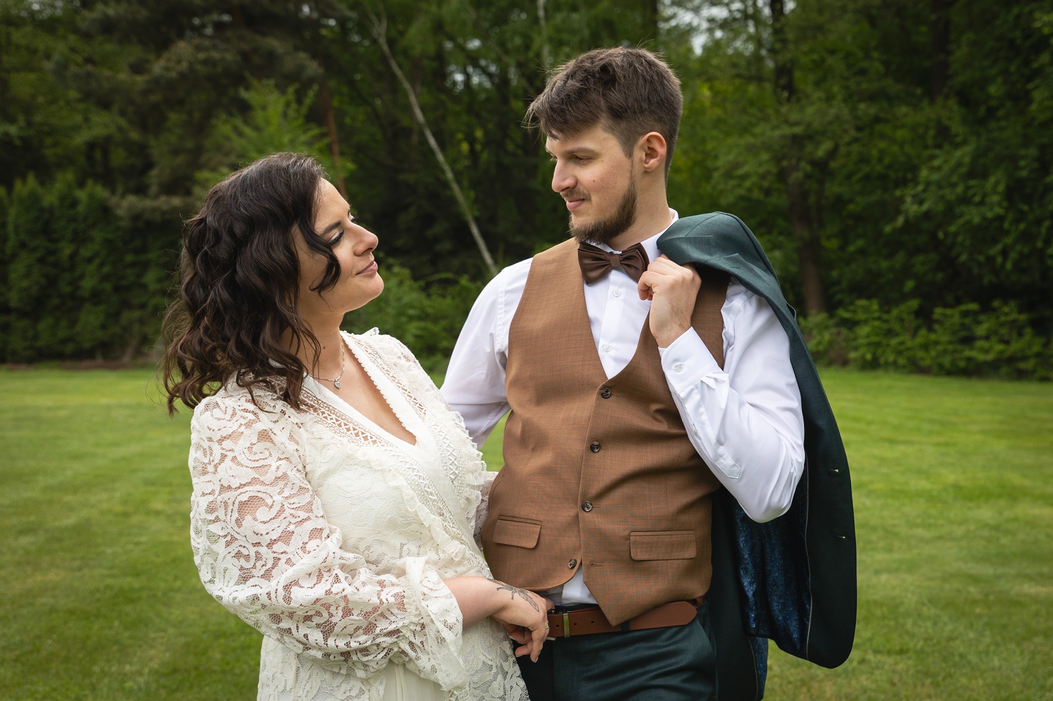 Uśmiechnięta para młoda w plenerze: panna młoda w koronkowej sukni spogląda na pana młodego w brązowej kamizelce i muszce, z marynarką przewieszoną przez ramię.