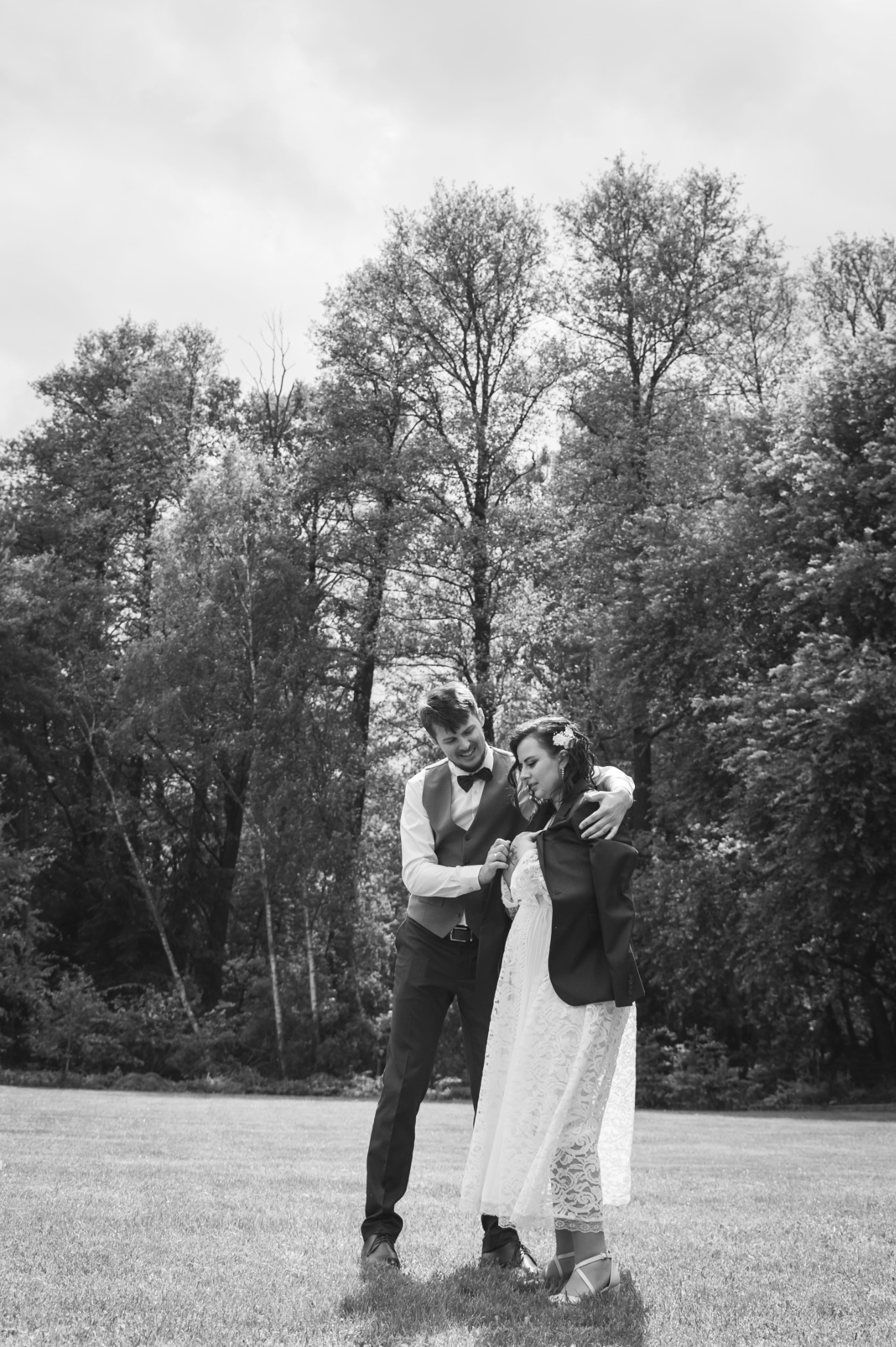 Czarno-białe zdjęcie pary młodej na tle lasu. Pan młody obejmuje pannę młodą, która poprawia marynarkę. Romantyczny nastrój w plenerze.