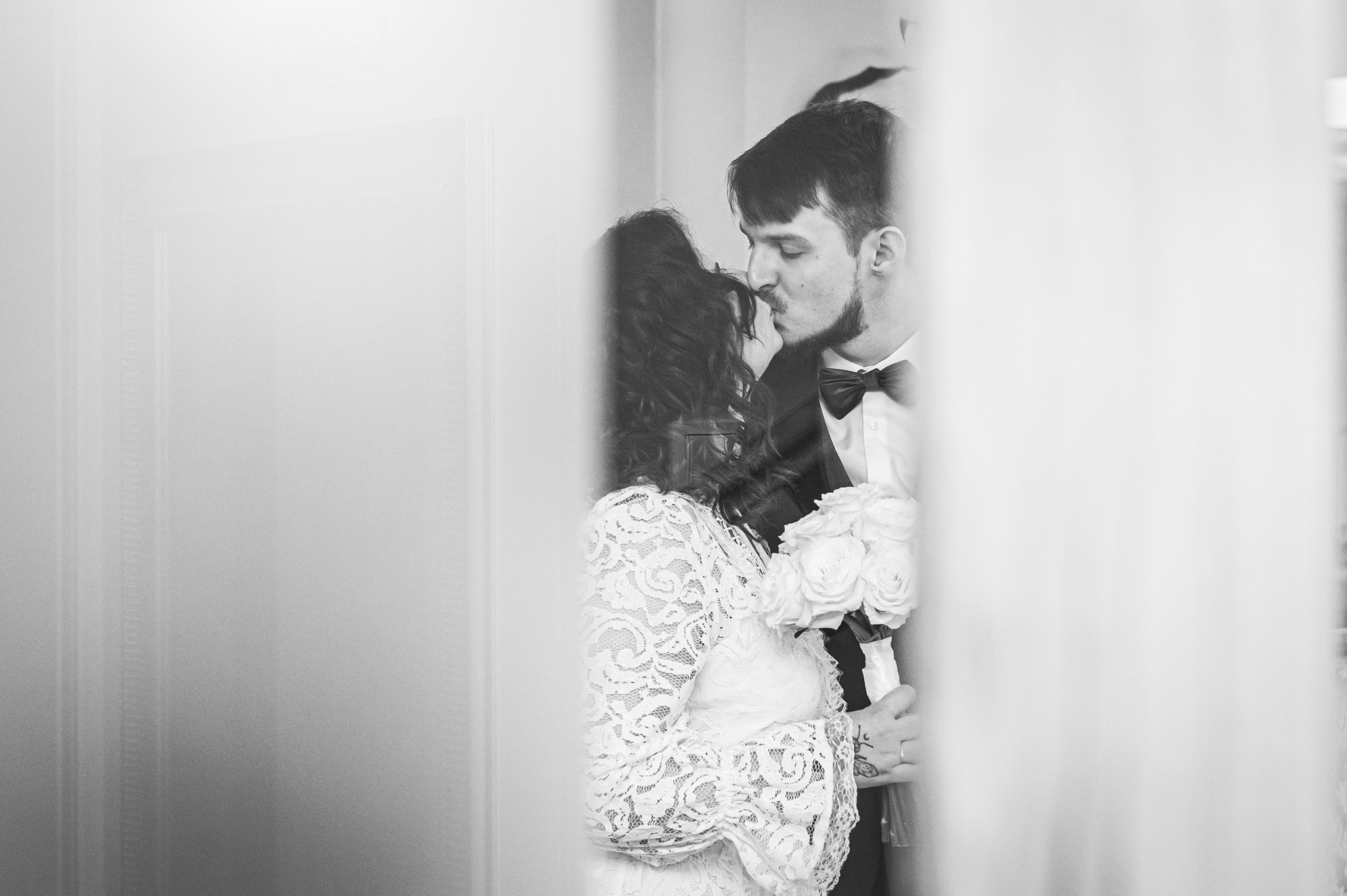 Czarno-białe zdjęcie pary młodej w zbliżeniu; panna młoda w koronkowej sukni trzyma bukiet, pan młody w muszce całuje ją w czoło, częściowo zasłonięci przez firankę.