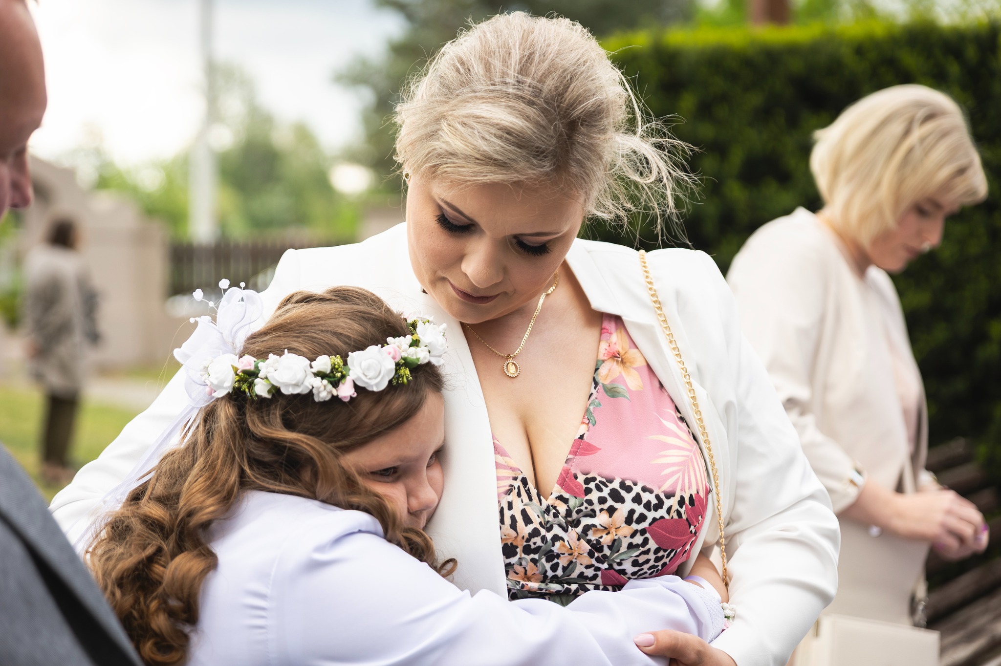 Wzruszające ujęcie: blond mama w białym żakiecie tuli zapłakaną dziewczynkę w wianku, na tle zieleni i jasnych budynków. Scena uchwycona z bliska, pełna emocji.