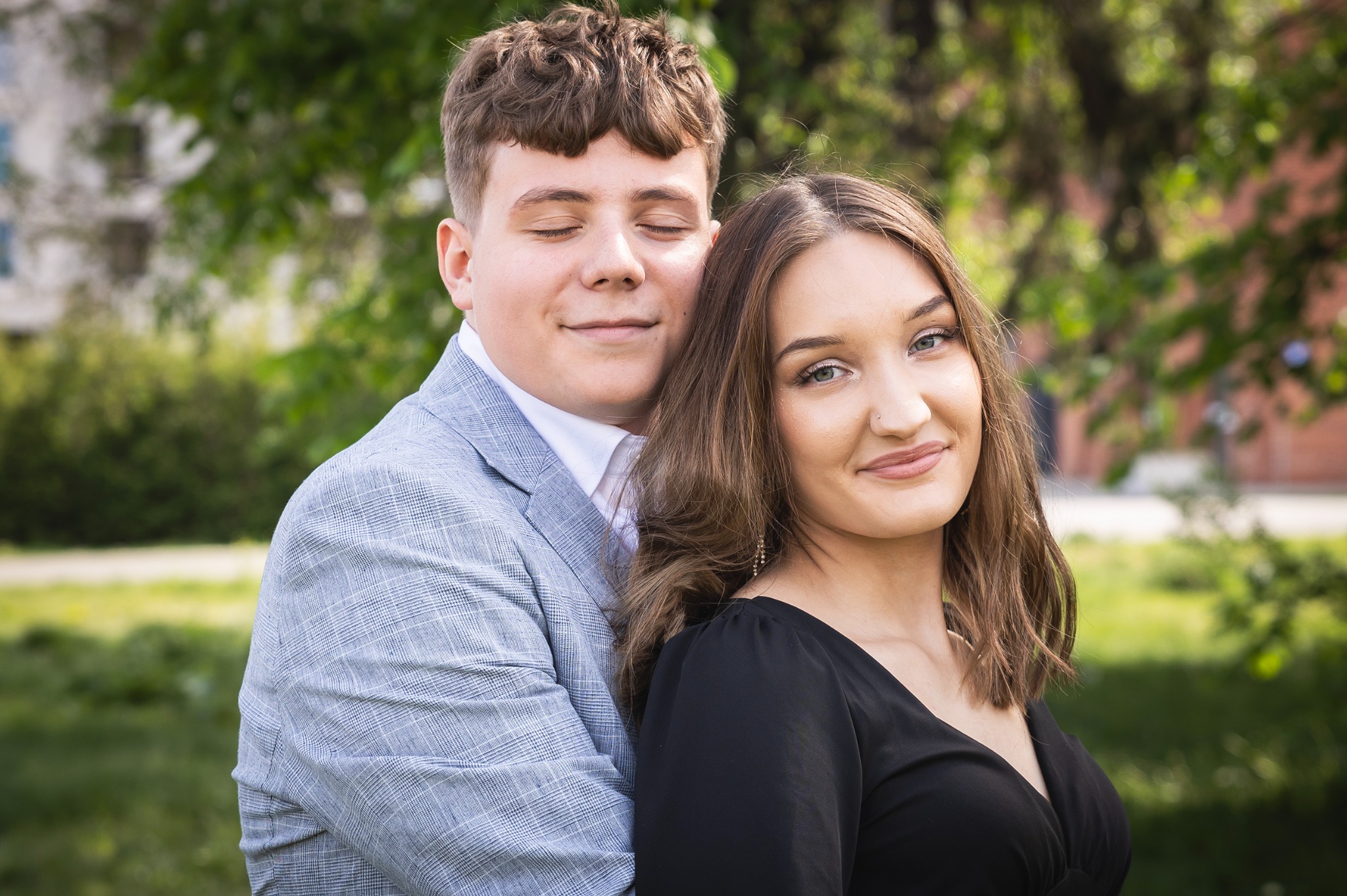 Uśmiechnięta para: mężczyzna w szarej marynarce obejmuje kobietę w czarnej sukience na tle zieleni. Mężczyzna ma zamknięte oczy, kobieta spogląda w obiektyw.